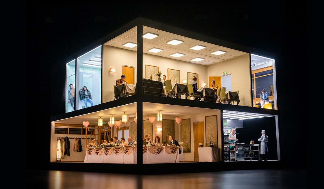 A two story building is set on stage. Inside we see people eating at tables in a restaurant, chefs in a kitchen and people in a bathroom. The lower floor is guests at a wedding reception dinner, there are heart balloons on the ceiling and guests are chatting.