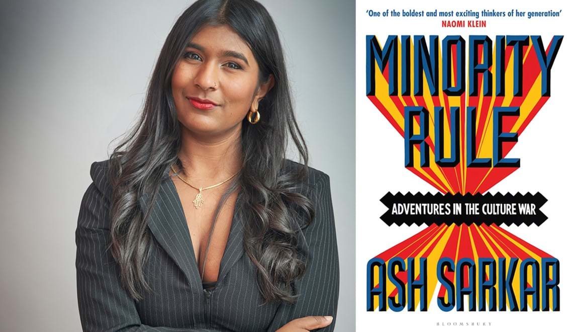 Ash wears a black blazer with gold jewellery. Ash has long wavy brown hair and is smiling.